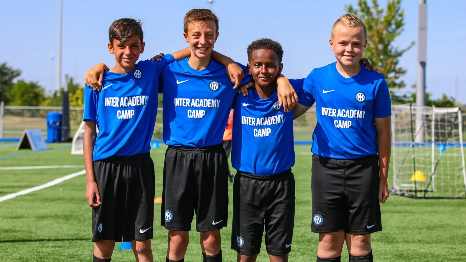 Four young football players in blue Inter Academy Camp jerseys and black shorts standing together on a green pitch.