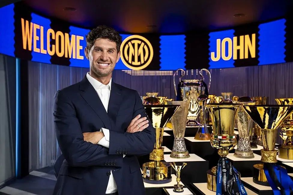 John Stones smiling in a navy suit standing in front of Inter Milan trophies and a Welcome John sign