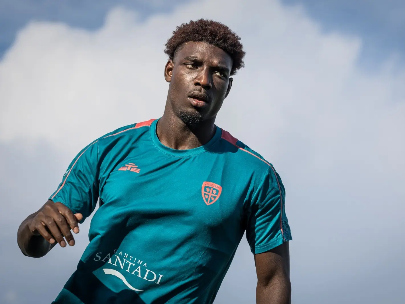 Michel Adopo wearing a teal Cagliari Calcio training shirt against a cloudy sky