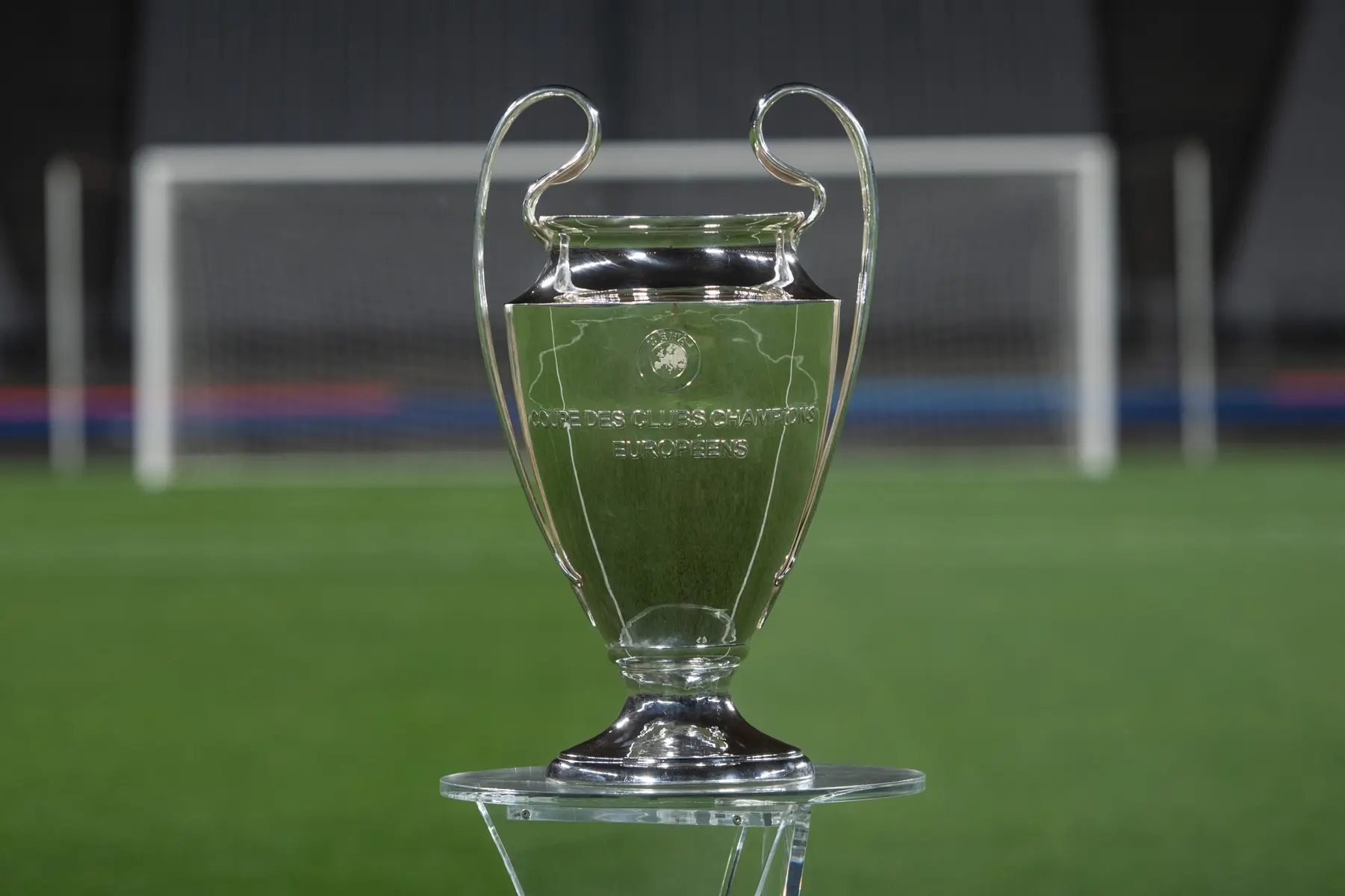 The silver UEFA Champions League trophy standing on a clear pedestal on a green football pitch